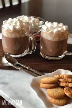 two mugs filled with hot chocolate and marshmallows on a plate next to cookies