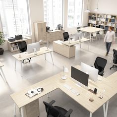 an office filled with desks and chairs in front of large windows, two people walking by