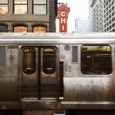 a silver train traveling past tall buildings on a city street in front of a neon sign