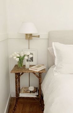 a small table with flowers on it next to a white bed and nightstand in a bedroom