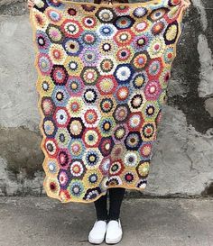 a woman is holding up a crocheted granny's afghan in front of a stone wall