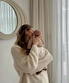 a woman holding a baby in her arms while standing next to a window with curtains