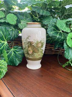 a white vase sitting on top of a wooden table next to green leaves and plants