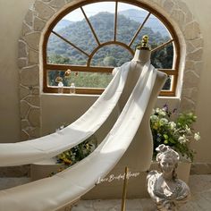 a mannequin draped in white fabric next to a window with mountains and flowers