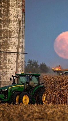 two tractors are driving through a field near a silo with a full moon in the background