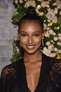 a woman in a black dress smiling at the camera with flowers behind her and a brick wall behind her