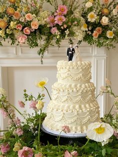 a wedding cake is surrounded by flowers and greenery with a bride and groom figurine on top