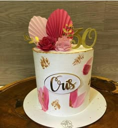 a white cake with pink flowers and gold lettering on the top is decorated with paper fans