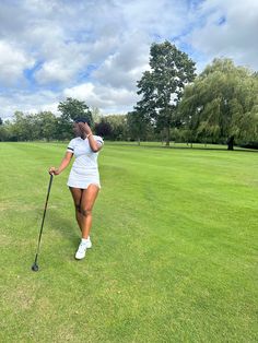 a woman walking across a lush green field holding a golf club and wearing a white dress