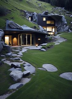 the house is surrounded by large rocks and green grass, with lights on in the windows