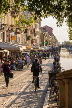 many people are walking and riding bikes on the sidewalk next to some water with buildings in the background