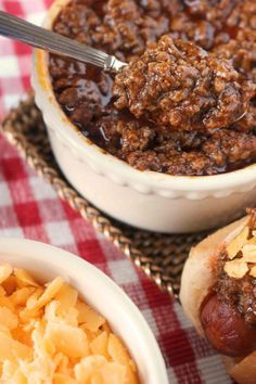 two hotdogs with chili and macaroni on a table next to a bowl of macaroni and cheese