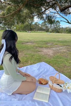 a woman sitting on a blanket with a picnic basket and book in front of her