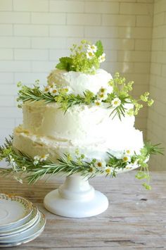 a three tiered cake with white frosting and greenery sits on a table next to plates