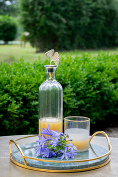 a glass bottle and some flowers on a table with a gold rimmed tray in front of bushes