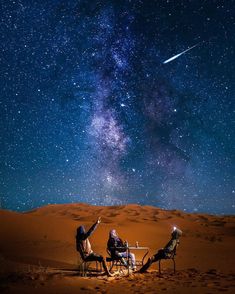 two people sitting in chairs on top of a sand dune under the night sky with stars