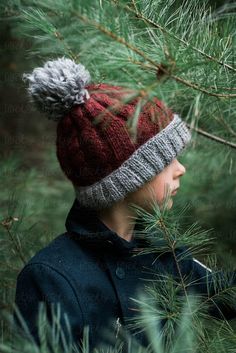 a young boy wearing a red and gray knitted hat standing next to a pine tree