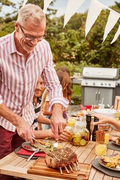 an older man cutting up food on top of a wooden table next to other people