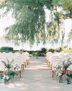 an outdoor ceremony setup with white chairs and greenery on the aisle, under a large willow tree