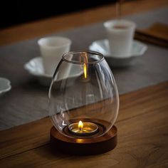 a lit candle in a glass vase on a wooden table with cups and saucers