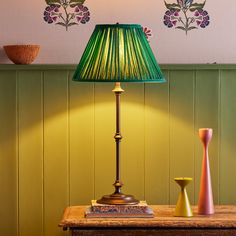 a green lamp on a table next to a book and vase with flowers painted on the wall behind it