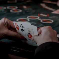 two hands holding playing cards in front of a black table with red and white numbers