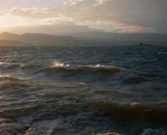 the sun is setting over the ocean with mountains in the backgrouund and clouds in the sky