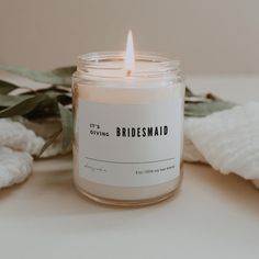 a white candle sitting on top of a table next to a towel and leaves with the words, it's giving bridesmaid