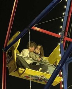 a man and woman sitting on a swing ride at night with lights in the background