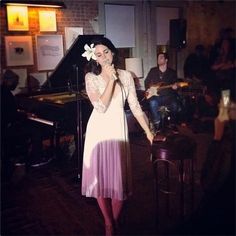 a woman singing into a microphone while standing in front of a piano and other people