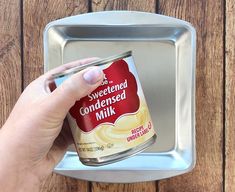 a hand is holding a can of sweetened condensed milk on a metal tray against a wooden wall