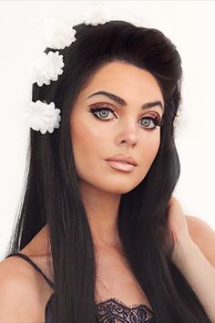 Jackie Wyers wearing a black wig with white flowers throughout the crown of the head. Her makeup is in the style of Priscilla Presley with thin dark brows, graphic liner, a tan and a frosty nude lip with a darker lip liner. Priscilla Presley 60s, 60s Makeup And Hair, 60s Makeup Tutorial, 70s Makeup Look, 70s Hair And Makeup, Jackie Wyers, Look Disco, Hippie Makeup, Disco Makeup