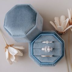 two engagement rings in a blue velvet ring box next to some flowers and buds on a white surface