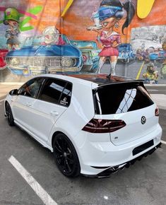 the back end of a white car parked in front of a wall with graffiti on it