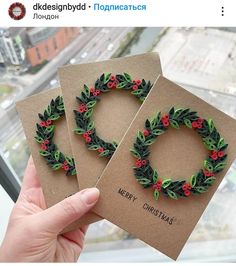 two handmade christmas cards with holly wreaths and red berries on them, held in front of a window