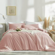 a bed with pink comforter and pillows in a white room next to a window