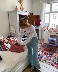 a woman standing on top of a bed next to a white dresser and window in a bedroom