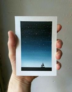 a person holding up a small card with a boat on the water and stars in the sky