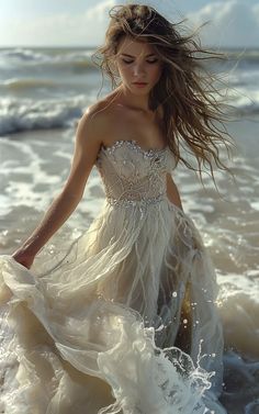 a woman in a white dress walking into the water at the beach with her hair blowing in the wind