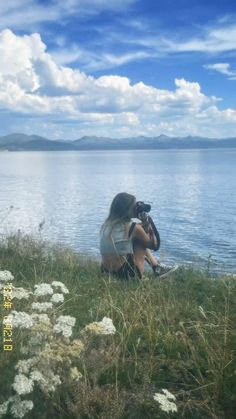 a woman sitting on top of a grass covered hillside next to the ocean with a camera