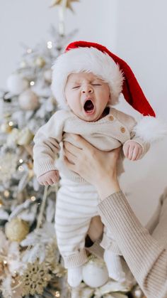 a woman holding a baby in front of a christmas tree wearing a santa claus hat