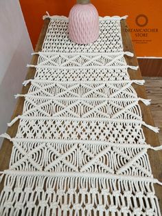 a white crocheted table runner with a pink vase on the top and an orange wall behind it