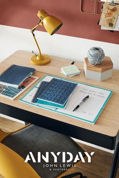 a desk with an open notebook and pen on it next to a lamp, books, and other items