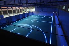 an indoor basketball court is lit up with blue lights in the dark, and it's surrounded by glass walls