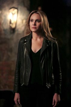 a woman in black leather jacket standing next to a wall with a clock on it