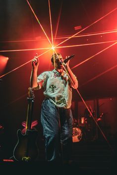 a man standing on stage holding a guitar and microphone in front of red light beams