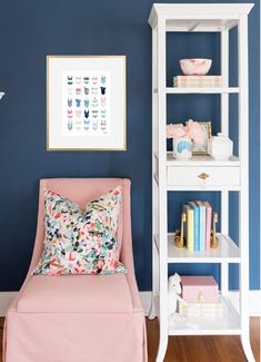 a pink chair sitting in front of a blue wall next to a white book shelf