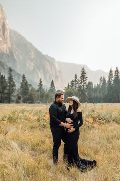 a pregnant couple standing in the grass with mountains in the background