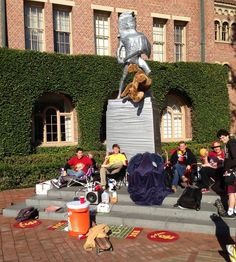 a group of people sitting on steps in front of a building with a statue of a man holding a teddy bear