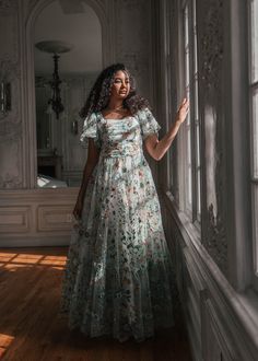 a woman in a long dress leaning against a wall with her hand on the window sill